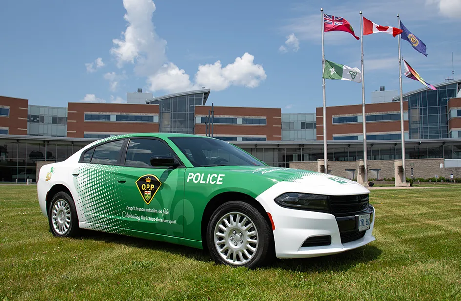 Voiture de police verte et blanche portant les armoiries de l’O.P.P. et le slogan « L’esprit franco-ontarien en fête / Celebrating the Franco-Ontarian spirit ». Derrière, un édifice et cinq drapeaux, dont franco-ontarien, Ontario, Canada et fierté LGBTQ.