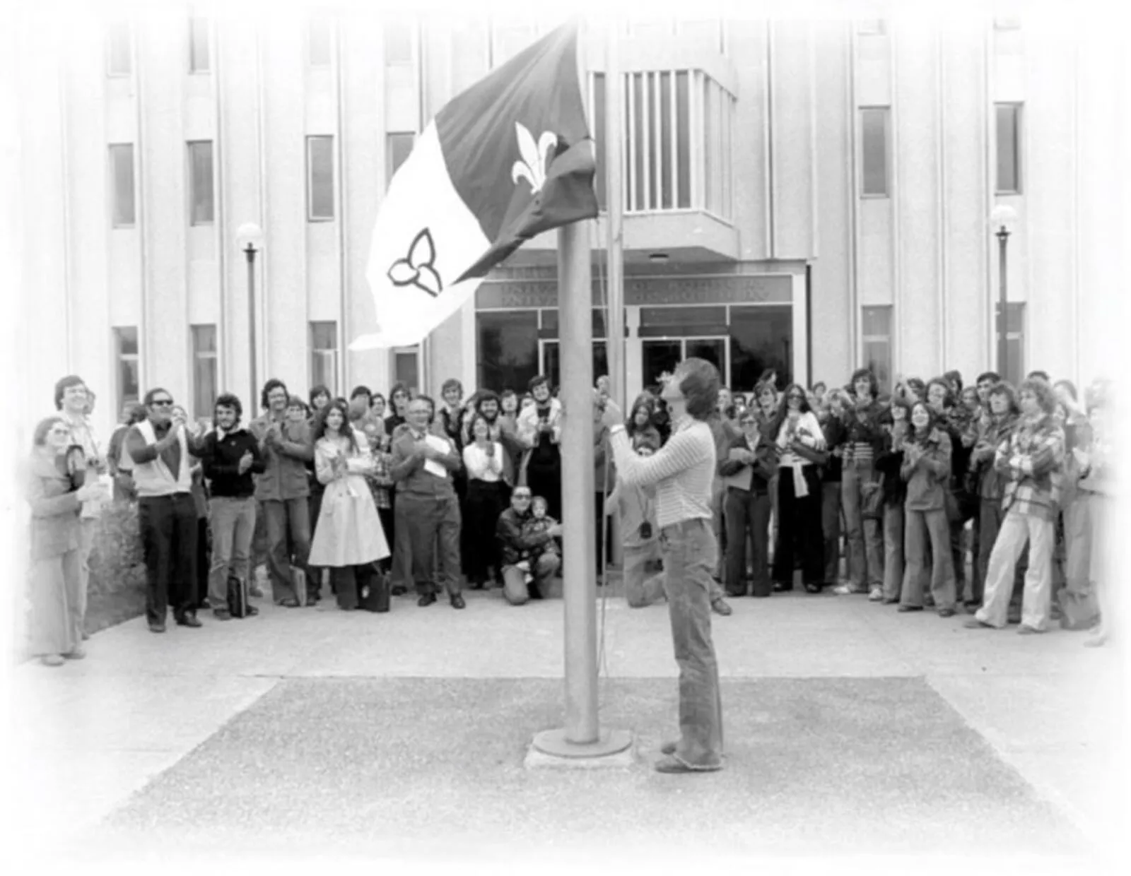 Photo en noir et blanc de Michel Dupuis en train de hisser le drapeau franco-ontarien devant l’Université de Sudbury, le 25 septembre 1975.