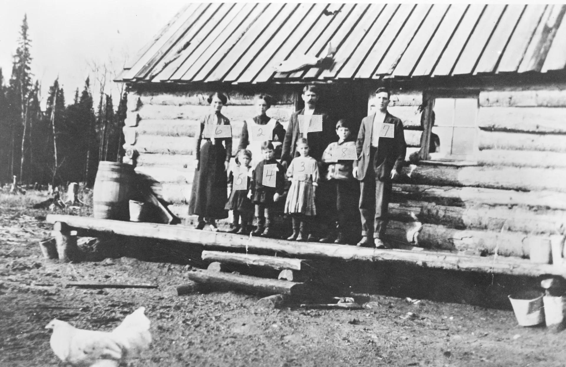 Photo en noir et blanc montrant une famille francophone sur le perron de sa maison en bois rond.