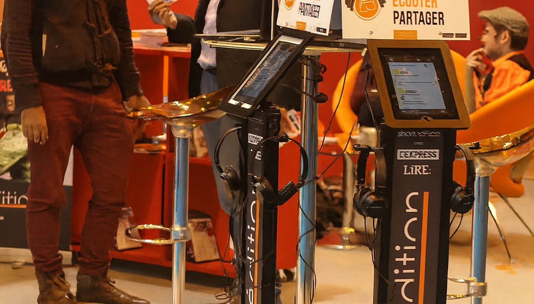 Dans un espace public ouvert, deux hommes discutent derrière des bornes Shortédition, semblables à des lutrins avec tablette électronique. Une affiche au-dessus, illustrant des écouteurs, porte le slogan : « Lire, écouter, partager ».