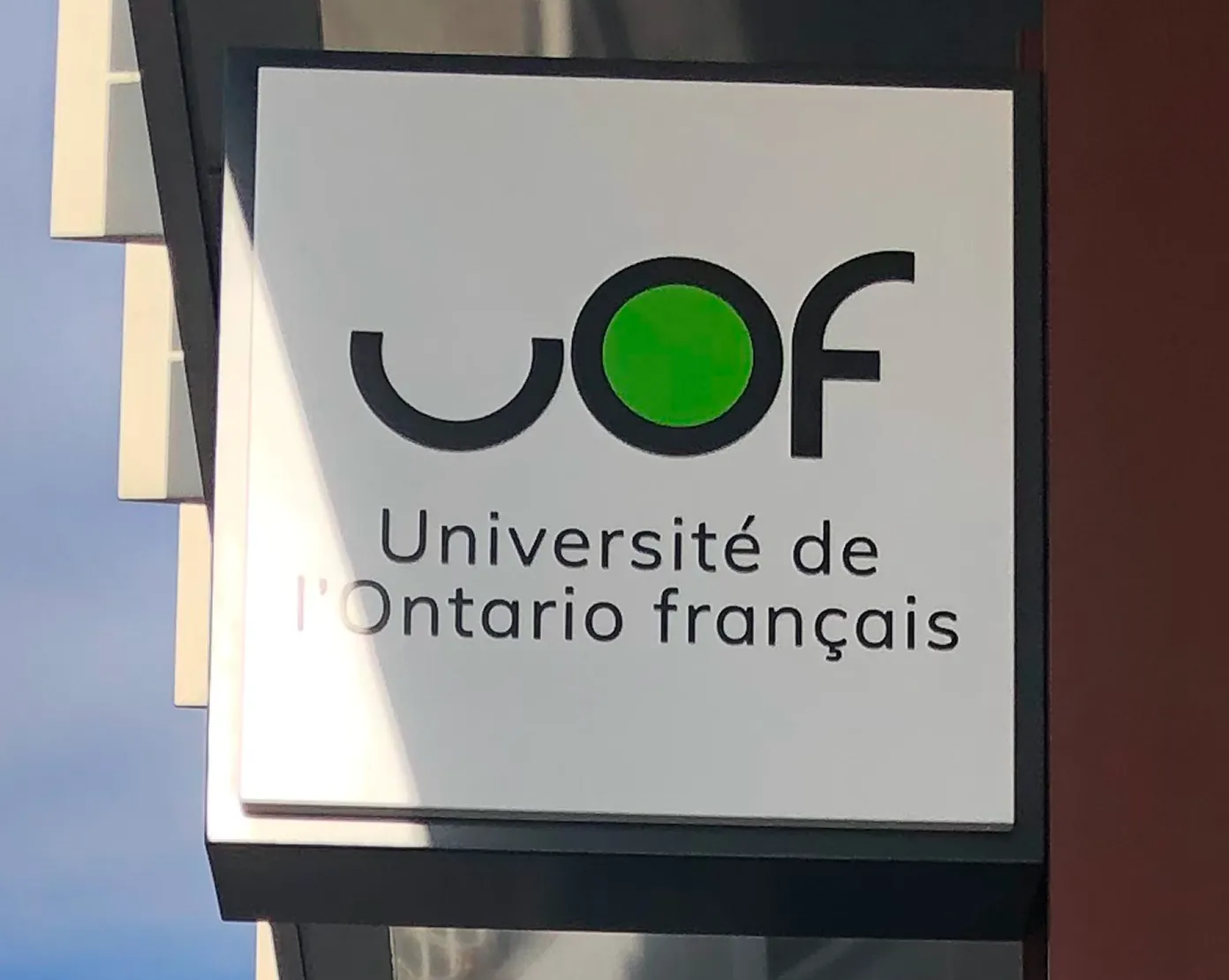 Logo de l’Université de l’Ontario français (UOF) sur un panneau blanc avec lettres noires et cercle vert, affiché sur la façade d’un bâtiment moderne.